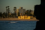 A beach scene at sunset with a yellow building near the shoreline, surrounded by tall trees and modern apartment buildings in the background. The ocean in the foreground is calm with a few visible waves, and there are people walking on the beach and in the water.