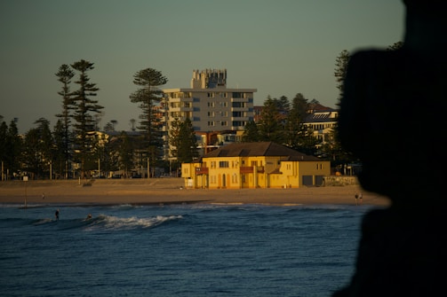 A beach scene at sunset with a yellow building near the shoreline, surrounded by tall trees and modern apartment buildings in the background. The ocean in the foreground is calm with a few visible waves, and there are people walking on the beach and in the water.