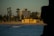 A beach scene at sunset with a yellow building near the shoreline, surrounded by tall trees and modern apartment buildings in the background. The ocean in the foreground is calm with a few visible waves, and there are people walking on the beach and in the water.
