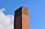 A tall brick chimney stands prominently against a clear blue sky. The brickwork is varying shades of red and brown, and the chimney has signs of weathering. Some clouds are visible in the background.