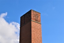 A tall brick chimney stands prominently against a clear blue sky. The brickwork is varying shades of red and brown, and the chimney has signs of weathering. Some clouds are visible in the background.