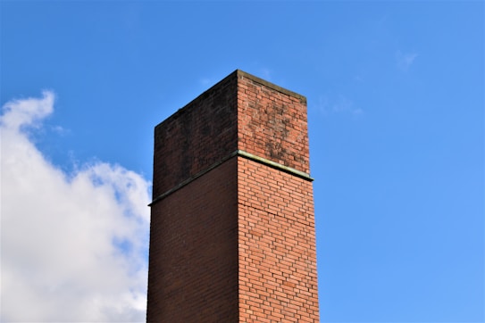 A tall brick chimney stands prominently against a clear blue sky. The brickwork is varying shades of red and brown, and the chimney has signs of weathering. Some clouds are visible in the background.