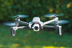 A drone with four propellers is captured in mid-flight against a blurred green background, indicating it is outdoors. The drone has a sleek, modern design with a camera integrated at its front.