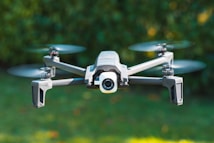 A drone with four propellers is captured in mid-flight against a blurred green background, indicating it is outdoors. The drone has a sleek, modern design with a camera integrated at its front.