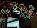 A vibrant urban scene featuring colorful neon artwork against a dark background. The neon outlines depict various playful images such as hands holding drinks, a person with a pizza slice, a space theme with a planet, and other abstract shapes. The phrase 'Let's Play Fashion' is prominently displayed in the center, highlighting a fashion-related theme. Below the neon art, silhouetted people gather in front of a lit structure with the same phrase.