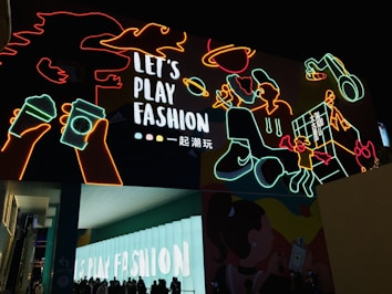 A vibrant urban scene featuring colorful neon artwork against a dark background. The neon outlines depict various playful images such as hands holding drinks, a person with a pizza slice, a space theme with a planet, and other abstract shapes. The phrase 'Let's Play Fashion' is prominently displayed in the center, highlighting a fashion-related theme. Below the neon art, silhouetted people gather in front of a lit structure with the same phrase.