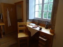 A cozy corner with a wooden desk and chair next to a window. The desk holds an open book, a ceramic mug, and a few pottery items on a white cloth. A wooden wardrobe stands nearby with a towel hanging from its door, and the window offers a view of greenery outside.