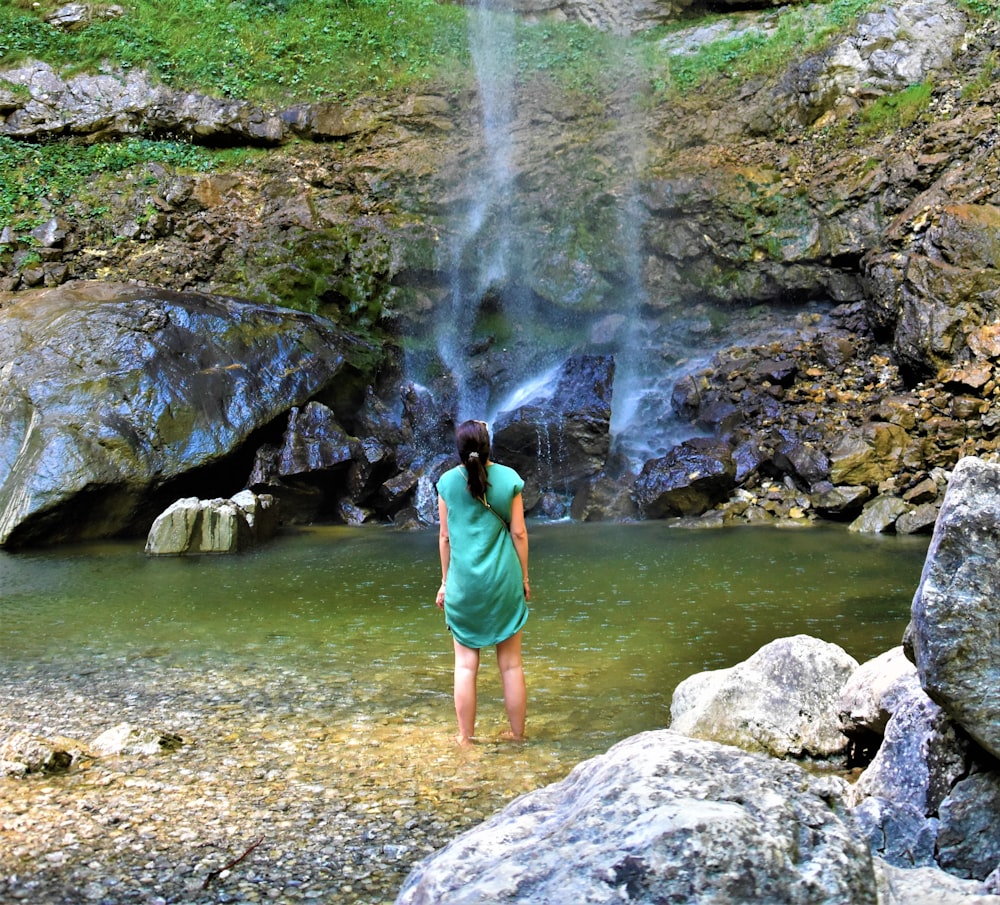 woman in teal tank top standing on rocky shore near waterfalls during daytime