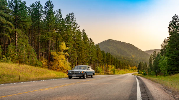 A vintage car driving through a forest road with autumn leaves creating a colorful backdrop.