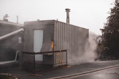 A large metal container with a white door is positioned on a slightly inclined surface. It is surrounded by mist or steam, creating a foggy effect. The container is secured with a metal fence adorned with yellow caution tape. Nearby structures are partially visible, including a rooftop and foliage.