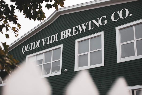 A green building features the text 'QUIDI VIDI BREWING CO.' in large white letters. The structure has several white-framed windows and is partially obscured by a white fence. Leaves are visible at the top of the image, suggesting nearby trees.