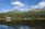 A tranquil mountain landscape with a dense evergreen forest reflected in a calm lake. A quaint wooden cabin with a gray roof sits nestled among the trees near the water’s edge. The backdrop features majestic mountain peaks under a bright blue sky with a few scattered clouds.