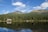 A tranquil mountain landscape with a dense evergreen forest reflected in a calm lake. A quaint wooden cabin with a gray roof sits nestled among the trees near the water’s edge. The backdrop features majestic mountain peaks under a bright blue sky with a few scattered clouds.