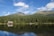 A tranquil mountain landscape with a dense evergreen forest reflected in a calm lake. A quaint wooden cabin with a gray roof sits nestled among the trees near the water’s edge. The backdrop features majestic mountain peaks under a bright blue sky with a few scattered clouds.