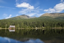 A tranquil mountain landscape with a dense evergreen forest reflected in a calm lake. A quaint wooden cabin with a gray roof sits nestled among the trees near the water&rsquo;s edge. The backdrop features majestic mountain peaks under a bright blue sky with a few scattered clouds.