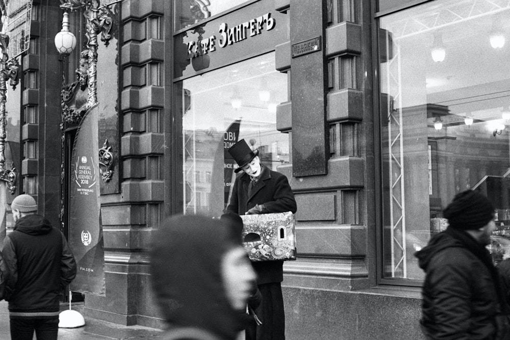 grayscale photo of man in black jacket and hat standing near building