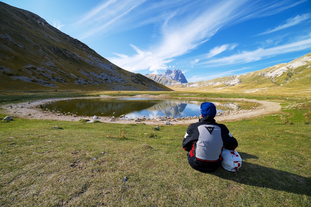 낮 동안 호수 근처의 푸른 잔디밭에 앉아 있는 빨간색과 검은색 재킷을 입은 사람