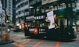 A green double-decker tram is driving through an urban street intersection. The tram is adorned with advertisements featuring text and a large image of a smartphone. Surrounding buildings are densely packed and typical of an urban environment. Neon signs and streetlights add to the vibrant city atmosphere.