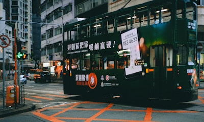 A green double-decker tram is driving through an urban street intersection. The tram is adorned with advertisements featuring text and a large image of a smartphone. Surrounding buildings are densely packed and typical of an urban environment. Neon signs and streetlights add to the vibrant city atmosphere.