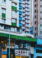 A vibrant urban scene featuring colorful high-rise buildings typical of a densely populated city. The architecture is varied with contrasting blocks of green, pink, blue, and white. In the foreground, a storefront sign reads 'Shing Fat Coconut Co.' indicating a commercial area. A construction crane is visible in the background, suggesting ongoing development or maintenance.