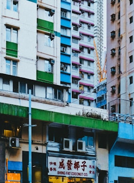 A vibrant urban scene featuring colorful high-rise buildings typical of a densely populated city. The architecture is varied with contrasting blocks of green, pink, blue, and white. In the foreground, a storefront sign reads 'Shing Fat Coconut Co.' indicating a commercial area. A construction crane is visible in the background, suggesting ongoing development or maintenance.