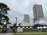 A weekend market setup in an outdoor urban setting with tall buildings in the background. Large umbrella-like structures provide shade over various stalls, some with white tents. Signs display information about the Greenfield Weekend Market. There are a few trees and a grassy area, with tables and chairs arranged for visitors.