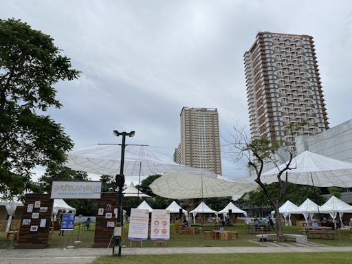 A weekend market setup in an outdoor urban setting with tall buildings in the background. Large umbrella-like structures provide shade over various stalls, some with white tents. Signs display information about the Greenfield Weekend Market. There are a few trees and a grassy area, with tables and chairs arranged for visitors.