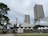 A weekend market setup in an outdoor urban setting with tall buildings in the background. Large umbrella-like structures provide shade over various stalls, some with white tents. Signs display information about the Greenfield Weekend Market. There are a few trees and a grassy area, with tables and chairs arranged for visitors.
