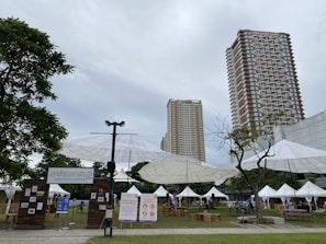 A weekend market setup in an outdoor urban setting with tall buildings in the background. Large umbrella-like structures provide shade over various stalls, some with white tents. Signs display information about the Greenfield Weekend Market. There are a few trees and a grassy area, with tables and chairs arranged for visitors.
