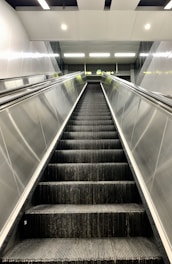 A modern, empty escalator in a well-lit, indoor environment. The escalator is flanked by metallic side panels and leads upwards to a landing area illuminated by overhead lights.
