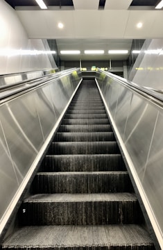 A modern, empty escalator in a well-lit, indoor environment. The escalator is flanked by metallic side panels and leads upwards to a landing area illuminated by overhead lights.