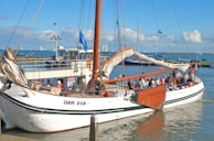 A sailboat named 'Carpe Diem' is docked in a harbor, filled with people enjoying a sunny day. The boat has white sails and wooden accents. In the background, other boats and a clear blue sky with some clouds are visible.