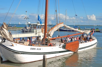 A sailboat named 'Carpe Diem' is docked in a harbor, filled with people enjoying a sunny day. The boat has white sails and wooden accents. In the background, other boats and a clear blue sky with some clouds are visible.