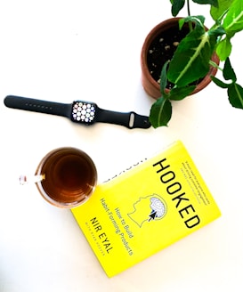 A pot with a green plant is placed on a white surface alongside a smartwatch, a glass of tea, and a yellow book titled 'Hooked' by Nir Eyal.
