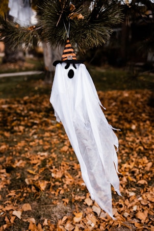 A ghost decoration with a white sheet drapes down, featuring a black, mouth-shaped cutout and eyepieces. It hangs from a tree branch, wearing a striped orange and black witch hat. The background is filled with dried leaves scattered on the ground, giving an autumn feel.