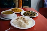 A red tabletop is set with three plates of food and a beverage can. One plate contains a mound of white rice topped with slices of poultry. Another plate holds a serving of yellow grains, possibly couscous or bulgur, accompanied by a green chili. The third dish appears to be a stir-fry with green vegetables and diced potatoes. A white bowl contains a greenish soup or stew.
