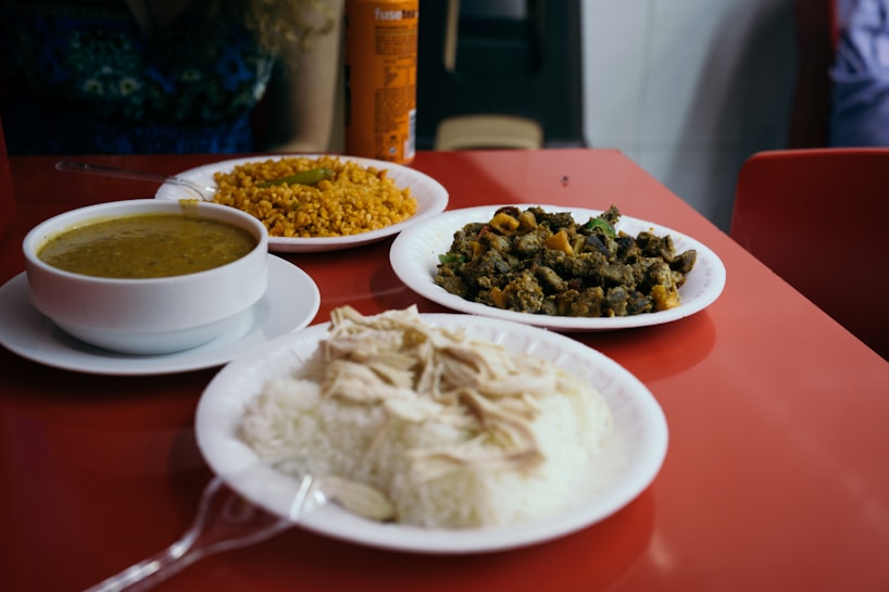 A red tabletop is set with three plates of food and a beverage can. One plate contains a mound of white rice topped with slices of poultry. Another plate holds a serving of yellow grains, possibly couscous or bulgur, accompanied by a green chili. The third dish appears to be a stir-fry with green vegetables and diced potatoes. A white bowl contains a greenish soup or stew.