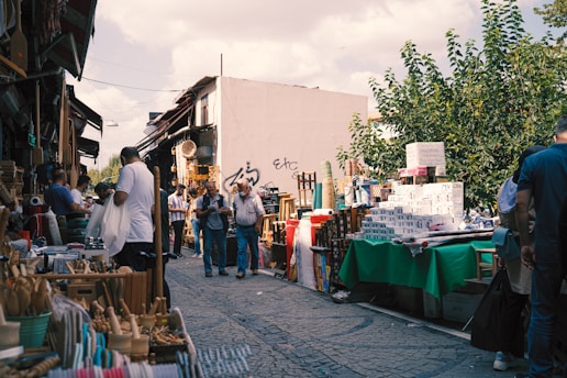 A bustling outdoor market scene with various stalls displaying a variety of items. People walk along the cobbled street, browsing and interacting with vendors. To the left, shelves are filled with kitchen utensils and wooden objects, while to the right, stacks of boxes and packaged goods are visible on tables covered with green cloth. The background shows a white building with graffiti and dense foliage from trees. The atmosphere is busy yet casual, typical of a lively market setting.