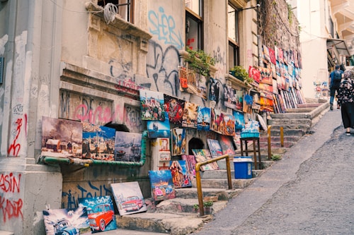 Numerous colorful paintings are displayed on the walls and ground along a narrow alley lined with steps. The setting gives an artistic and vibrant feel, featuring various landscapes, vintage cars, and city scenes. The walls show graffiti and climbing plants, and a few people are seen walking up the alley.