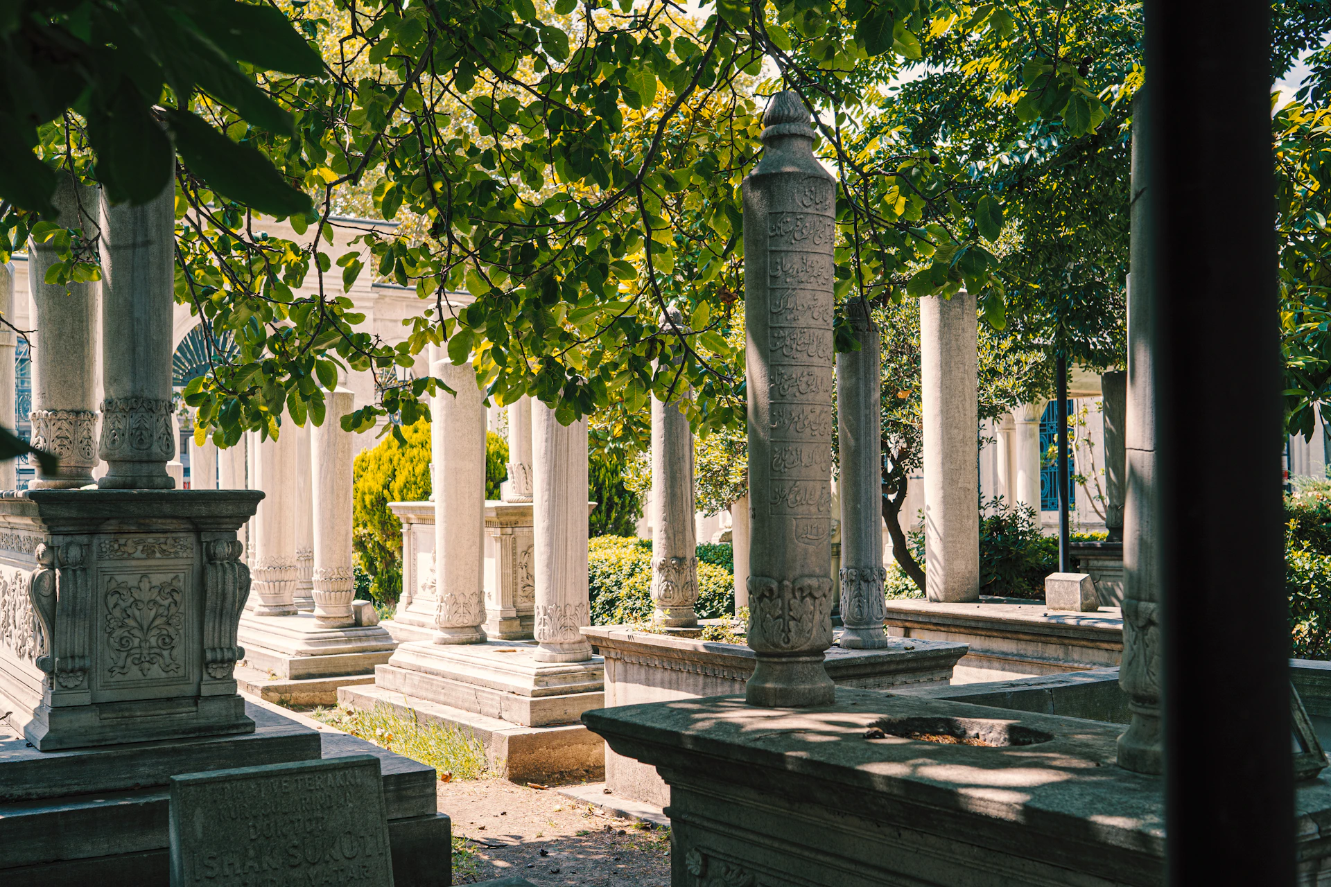 Columna decorativa de concreto con detalles grecorromanos en un jardín soleado, rodeada de plantas verdes.