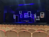 An empty theater with rows of light-colored wooden seats facing a dimly lit stage. The stage features several illuminated rectangular frames on the back wall, creating a modern and artistic backdrop. A bed with a blanket sprawled on it and a small table with various objects are also visible on the stage. The predominant lighting is blue, giving the scene a dramatic atmosphere.