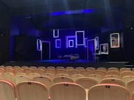 An empty theater with rows of light-colored wooden seats facing a dimly lit stage. The stage features several illuminated rectangular frames on the back wall, creating a modern and artistic backdrop. A bed with a blanket sprawled on it and a small table with various objects are also visible on the stage. The predominant lighting is blue, giving the scene a dramatic atmosphere.