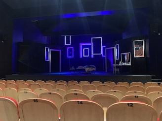 An empty theater with rows of light-colored wooden seats facing a dimly lit stage. The stage features several illuminated rectangular frames on the back wall, creating a modern and artistic backdrop. A bed with a blanket sprawled on it and a small table with various objects are also visible on the stage. The predominant lighting is blue, giving the scene a dramatic atmosphere.