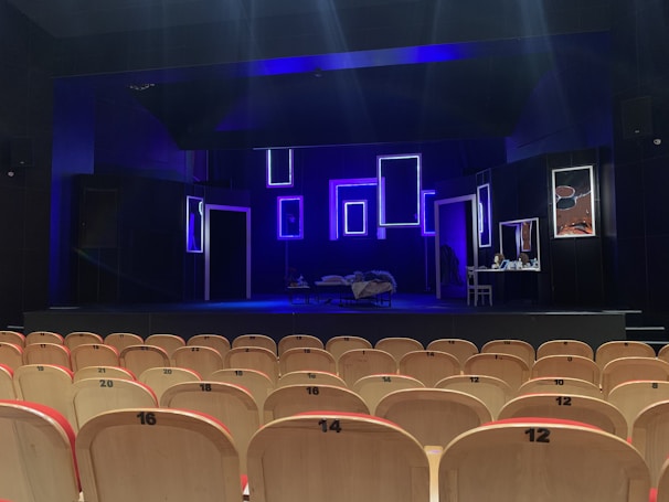 An empty theater with rows of light-colored wooden seats facing a dimly lit stage. The stage features several illuminated rectangular frames on the back wall, creating a modern and artistic backdrop. A bed with a blanket sprawled on it and a small table with various objects are also visible on the stage. The predominant lighting is blue, giving the scene a dramatic atmosphere.