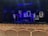 An empty theater with rows of light-colored wooden seats facing a dimly lit stage. The stage features several illuminated rectangular frames on the back wall, creating a modern and artistic backdrop. A bed with a blanket sprawled on it and a small table with various objects are also visible on the stage. The predominant lighting is blue, giving the scene a dramatic atmosphere.