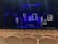 An empty theater with rows of light-colored wooden seats facing a dimly lit stage. The stage features several illuminated rectangular frames on the back wall, creating a modern and artistic backdrop. A bed with a blanket sprawled on it and a small table with various objects are also visible on the stage. The predominant lighting is blue, giving the scene a dramatic atmosphere.