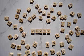 A collection of Scrabble tiles scattered randomly on a marble surface with the word 'CHAOS' spelled out in the center. The tiles are cream-colored with black letters, and the lighting creates shadows that add depth to the image.