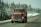 A red semi-truck with visible exhaust pipes is driving down a snowy road lined with evergreen trees. The truck features a message in white text on the front. The overcast sky and snow on the ground create a wintry atmosphere.