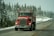 A red semi-truck with visible exhaust pipes is driving down a snowy road lined with evergreen trees. The truck features a message in white text on the front. The overcast sky and snow on the ground create a wintry atmosphere.