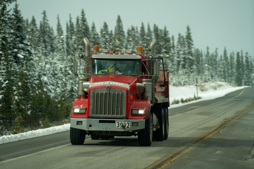 A red semi-truck with visible exhaust pipes is driving down a snowy road lined with evergreen trees. The truck features a message in white text on the front. The overcast sky and snow on the ground create a wintry atmosphere.